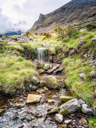 A small waterfall cascades over rocks and vegetation in a picturesque landscape. This is along the popular hiking trail of Carrauntoohill in County Kerry, Ireland, with a mountain in the background.の写真素材