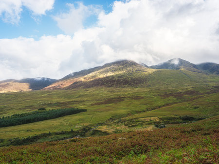 Green hills and mountains rise in the distance, partly covered by low-hanging clouds on Carrauntoohil walkway. Sunlight shines on the slopes in County Kerry, Ireland.の写真素材