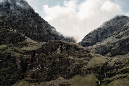 Fog drifts over rocky, green terrain leading up to the peak of Carrauntoohill in County Kerry, Ireland. A small waterfall trickles down the rugged mountain face.の写真素材