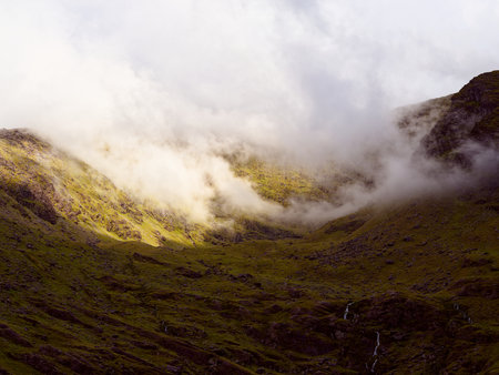 Misty clouds drift through the valley of Carrauntoohill. Low-hanging clouds obscure the mountains peak, adding to the dramatic atmosphere of the Irish landscape.の写真素材