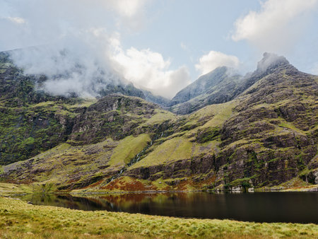 The lake is calm and reflects the mountains of Carrauntoohil in Ireland. On a dull day, fog and clouds make the mountain difficult to see.の写真素材