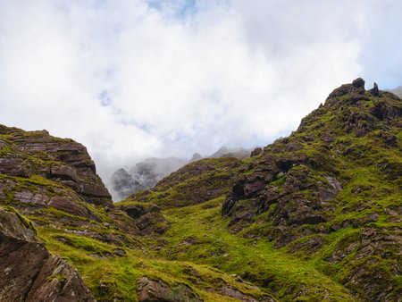 The view along the Carrauntoohill walkway is of a rugged, green landscape. On an overcast day, mist and clouds cover the higher peaks, making the cliffs and vibrant plants below stand out.の写真素材