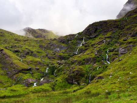 This vista showcases the lush, green, rolling landscape of Carrauntoohil, Ireland, with several small streams trickling down the hillside after rainfall on a cloudy day.の写真素材