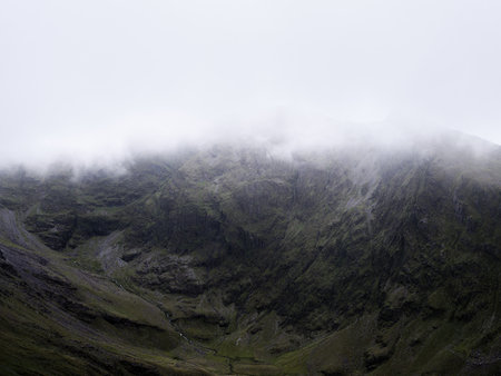 A mountain face is obscured by low-hanging fog or mist. Green slopes and craggy rock formations create a dramatic texture under a white sky.の写真素材