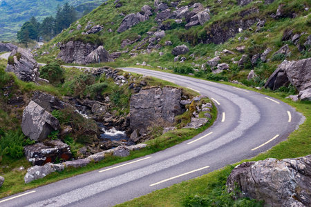 An asphalt road winds through a lush, rocky mountain pass. A small stream flows alongside the road in the Gap of Dunloe in County Kerry, Ireland.の写真素材