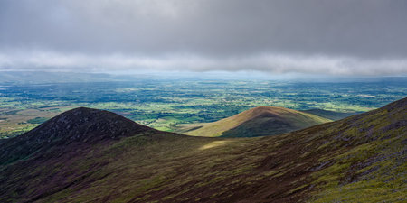 A view of the countryside in Ireland, with rolling hills and green valleys, as seen from the top of the Coomloughra Horseshoe, with a cloudy sky in the background.の写真素材