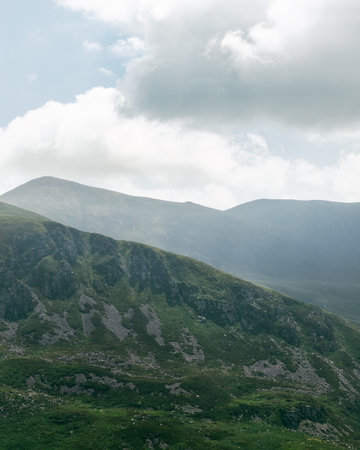 A scenic view captures the rugged green mountains in the Gap of Dunloe, Ireland. Clouds fill the...