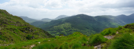 Rolling green hills and cloudy skies can be seen across the Gap of Dunloe in County Kerry, Ireland....
