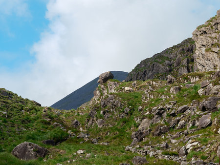 This showcases the rocky terrain and lush greenery of the Gap of Dunloe in Ireland. A mountain rises in the background under a partly cloudy sky.の写真素材