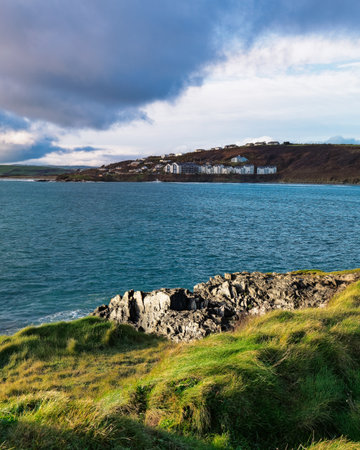 A beautiful view of Inchydoney Island in Ireland. The green grassy cliffs meet the blue water under a partly cloudy sky. Buildings sit atop the distant hill.の写真素材