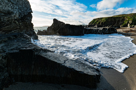 On a sunny day, waves from the ocean gently wash up on the black sand beach at Sheep Cove in Ireland, among the rocks and cliffs.の写真素材
