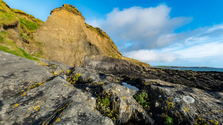 A rocky coastline features layered rock formations and green grass. A pool of water sits on the rocks, with the Atlantic Ocean and sky visible in the distance in West Cork.の写真素材