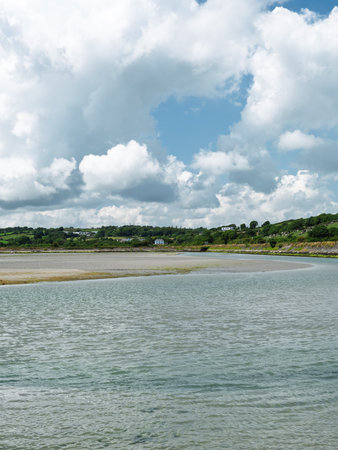 Expansive water stretches towards a sandy shore, framed by rolling green hills and a serene, cloud-dotted sky.の写真素材