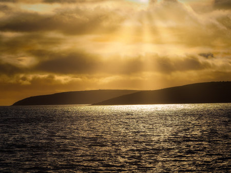 Golden sunlight filters through clouds and illuminates the ocean surface. Dark land masses are visible in the distance against the warm, colorful sky.の写真素材