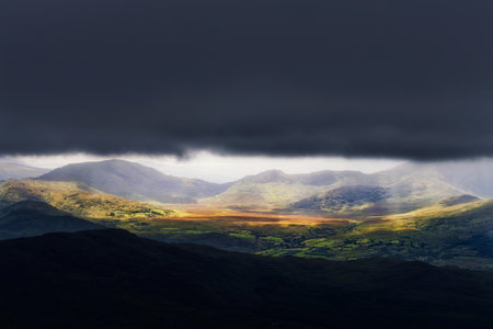 A picturesque view in Killarney National Park, Ireland. Sunlight breaks through a dark, stormy sky,...