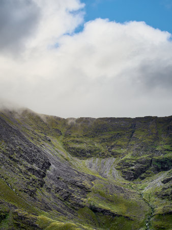 Clouds hover over a green and rocky mountainside, creating a scene of natural beauty and...