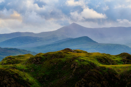 Green hills sit in the foreground, leading back to blue layered mountains. Clouds cover the sky above National Park during the daytime.の写真素材