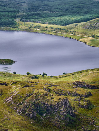 A lake stretches out surrounded by green hills and trees. Rocks form a terrace in the foreground. The day appears cloudy.の写真素材