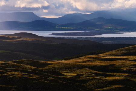 The hillsides glow in the soft light. Silhouetted mountains stand in the distance. Forests fill the valleys below. Clouds soften the sky above the mountain peaks.の写真素材