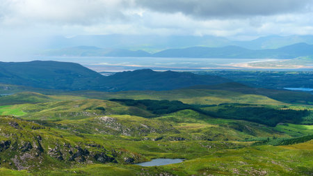 Green hills and forests meet a blue lake. Mountains fade into a bright, cloudy horizon.