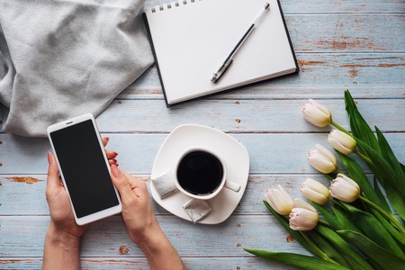 bouquet of white tulips with a Cup of coffee and a smartphone in womens hand and an empty notebook...