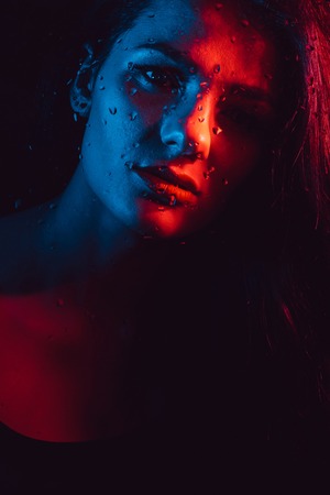 Sensual portrait of beautiful girl behind glass with raindrops with red blue lightingの写真素材