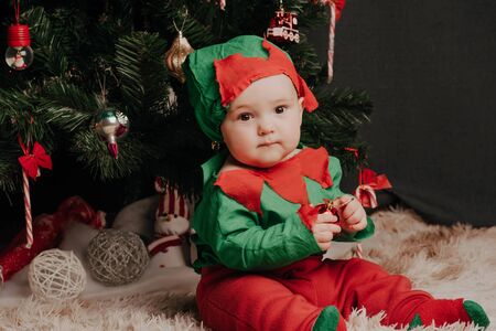 little boy in elf costume sits under a Christmas tree with balls