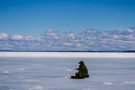 man fisherman with a fishing rod sitting and fishing on the ice in winterの写真素材