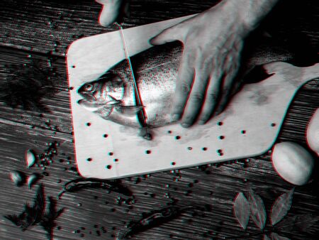 man cuts a trout with a knife on a Board on a wooden table. Fresh raw fish. Black and white photo with 3D glitch effectの写真素材
