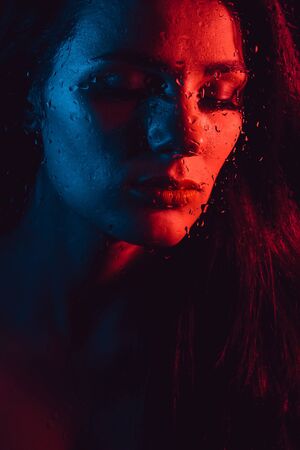 Sensual portrait of beautiful girl behind glass with raindrops with red blue lightingの写真素材