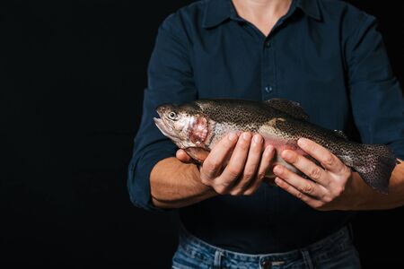 Studio shot of stylish young bearded fisherman in yellow raincoat and red hat looking in shock with jaw dropped, holding big sea-water fresh-caught fish in both hands, surprised with fine catchの写真素材