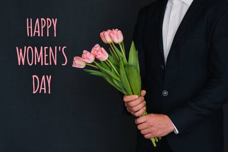 Bouquet of pink flowers of tulips in men's hands in a suit. Greeting card with the inscription Happy women's dayの写真素材