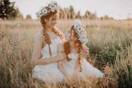 Happy mother and daughter are smiling and hugging in the field in summer in white dresses with braids and floral wreathsの写真素材