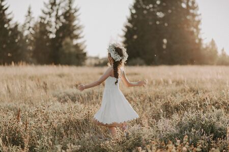 happy joyful young girl in a white dress and with a flower wreath on her head in the field in the summerの写真素材