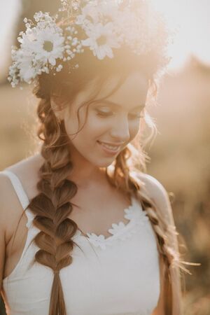 portrait of smiling girl in white dress with floral wreath and braids in summer at sunset in the fieldの写真素材