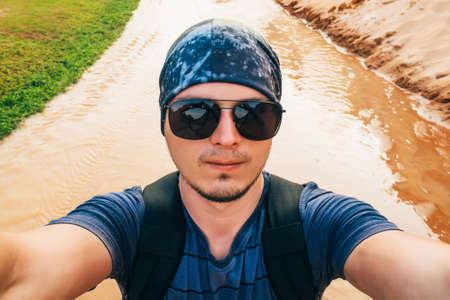man on a hike with a backpack takes a selfie on camera face in sunglasses and a bandana while...