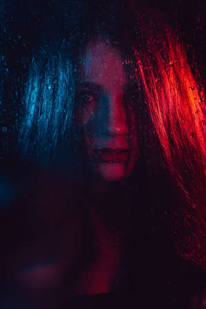 Sensual portrait of a beautiful attractive girl looking through the glass with raindropsの写真素材