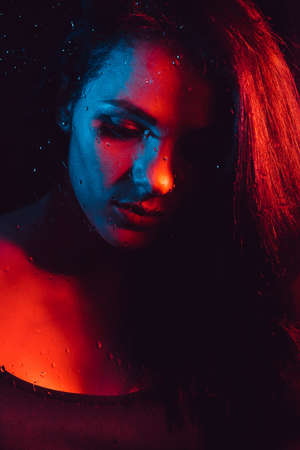Sensual portrait of beautiful girl behind glass with raindrops with red blue lightingの写真素材