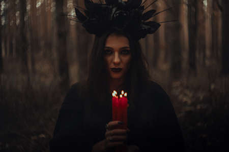witch in a black costume holds candles in a dark forest