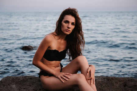 Sexy young girl in black swimsuit sitting on the rocks by the sea in summerの写真素材