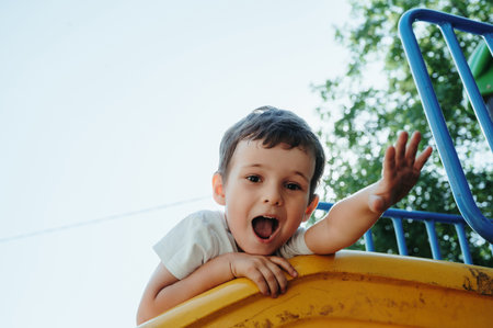 happy preschooler boy playing on slide on playground in summerの写真素材