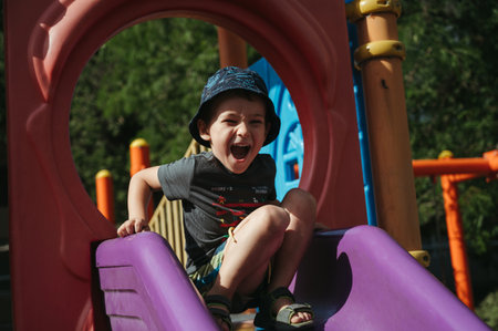 happy kindergarten boy rides a slide on playground in summerの写真素材