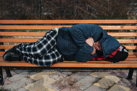 homeless dirty elderly old Caucasian man lies sleeping on park bench in autumnの写真素材