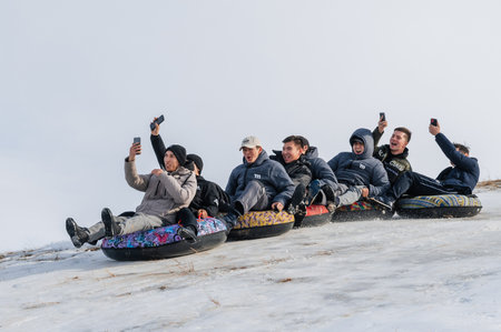 TAU SAMALY, KAZAKHSTAN - DECEMBER 23, 2023: happy Kazakhs friends tubing down a slide in winter with snow at Tau Samaly recreation center in Kazakhstanのeditorial素材