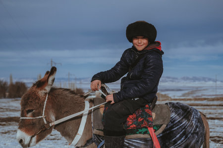 TAU SAMALY, KAZAKHSTAN - DECEMBER 23, 2023: Kazakh boy riding a donkey in winter in field near Tau Samaly in Kazakhstanのeditorial素材