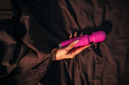 Pink silicone vibrator for masturbation in a female hand. A womans hand under blanket holds a sex toy for orgasm on bed in the bedroomの写真素材