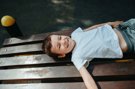 happy baby boy is lying on bench on playground in summerの写真素材
