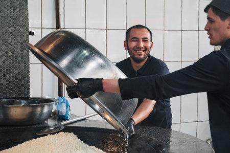 happy male chefs prepare rice for Uzbek pilaf in a cauldron in the kitchen of restaurant Central...