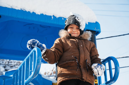 A toddler in a brown jacket and black hat is happily playing in the freezing snow in a public space, enjoying leisure fun in the winterの写真素材