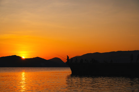 Silhouettes of fishermen with fishing rods fishing on pier in sea in evening at sunset in summerの写真素材